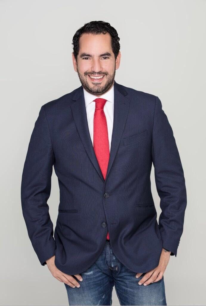 Rolando González de pie, sonriendo, con saco azul y corbata roja